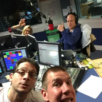 Two men pulling faces in a radio studio while presenters record behind them