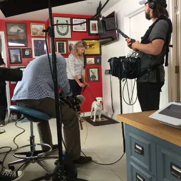 A film crew shooting an interview in a room hung with pictures, a dog watching from the floor