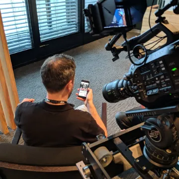 A camera operator filming a seated interviewee beside a window