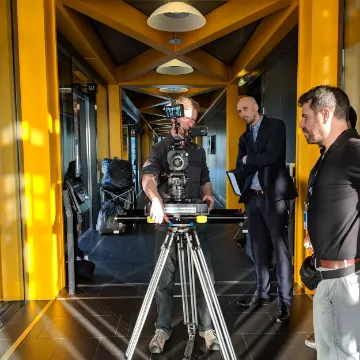 A camera on a tripod filming two men talking in a yellow-lit corridor
