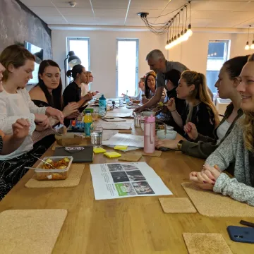 A long table of people in a workshop, papers and drinks spread across it