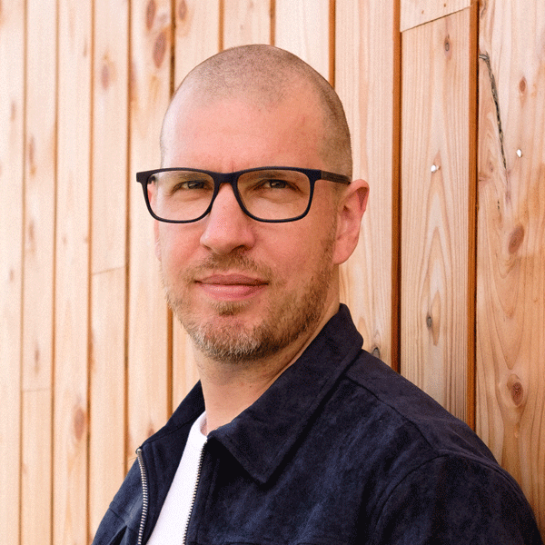 Portrait of Olly Whitman in glasses and a dark jacket against a timber-clad wall