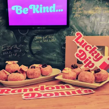 A counter of donuts under a “Random Donuts, Lucky Dip” sign, beside a screen reading “Be kind…”